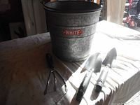 Galvanized White metal pail and Husky garden hand tools on a cloth surface, showing front bucket logo and tool handles.
