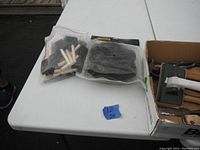 Photo showing bag of new foam brushes and new black knee pads in plastic bags on white table next to box of tools