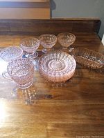 Set of pink depression glass including 6 plates, 4 footed small bowls, cream pitcher, and oblong dish on wooden surface.