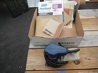 Blue Ryobi palm sander with attached dust bag and its power cord coiled beside a box containing various sheets of sandpaper.