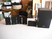 Sony surround sound speaker set including four satellite speakers, center speaker, and subwoofer, displayed on white table.