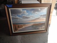 Framed beach picture showing sandy pathway, ocean, sky with clouds, and wooden fencing.