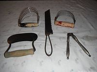 Four vintage kitchen tools laid out on a fabric surface: two wooden-handled dough cutters/pastry blenders, one metal bread slicer with scalloped blade, and metal garlic press.