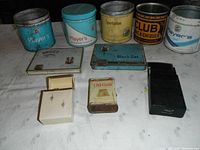 Group photo of seven vintage tobacco tins plus a small white tin labeled 'Sweet Vapa' and a black plastic case with Player's branding. Various colors and prints, mostly metal tins.