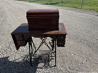 Full view of the closed wooden cabinet sewing machine on cast iron treadle stand outdoors on gravel.