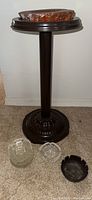 Full view of dark brown ashtray stand with decorative base, stone ashtray on top, and three glass ashtrays in front on carpet.