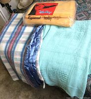 Three blankets displayed: plaid wool blend with satin border, mint green cotton woven, and packaged orange Edmond polyester