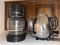 Black Proctor-Silex drip coffee maker with glass carafe and cord, shown next to a silver GE electric percolator kettle on a white surface with wooden backdrop.