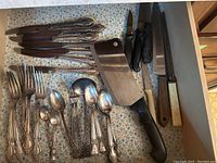 Overview of assorted cutlery and cutting utensils arranged in a drawer on floral liner, includes knives, forks, spoons, cleaver, sharpening tool.