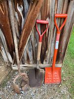 Photo showing three long-handled tools: two metal shovels with wooden handles and a manual post hole digger with an orange plastic handle.