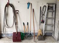 Wide view of the lot including hose, snow shovel, watering can, pruning shears, rake, garden fork, broom, metal dustpan, step ladder, and jumper cables.