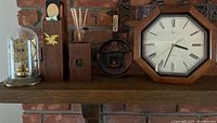 Shelf display of six mantel decor items against brick background: brass anniversary clock under glass dome, wooden match containers, decorative thermometer with fireplace carving, and wood framed wall clock.