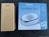 Both product boxes side by side on black surface showing front of Homemedics SoundSpa Ultra and Saje Aroma Roam diffuser.