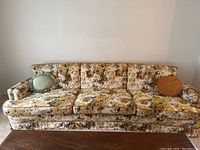 Full view of the three-seat retro sofa showing floral upholstery in yellow, gold, green and cream, with a mint green and burnt orange round pillow on either end.