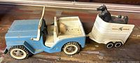 Side view of blue metal jeep attached to white horse trailer with two black plastic horses inside.
