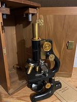Full view of black metal microscope with brass eyepiece and adjustment knobs inside wooden case with open door.