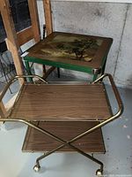 Photo of a vintage square card table with painted scenery on top and a brass/gold metal drinks cart with two shelves below.