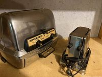 Photo showing both vintage toasters side by side on a wooden surface against a wall background. Larger toaster has drop down sides and control panel, smaller is General Electric toaster with knobs and cord.
