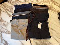 Overview of eight pairs of men's casual pants including khaki, corduroy, navy, gray, black, and brown colors, plus two brown leather belts on a marble surface.