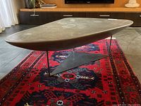 Side angle view showing the triangular fibreglass top with stone-like texture and the black metal base with triangular plate support, placed on a red patterned rug in a modern room.