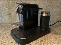 Front side view of the black Nespresso espresso machine showing drip tray and milk frother attached on the right side.