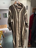 Front view of the brown and cream striped handwoven wool jalaba poncho style jacket hanging on a wooden hanger in a well-lit indoor area.