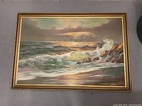 Framed painting showing waves and rocky shore under sunset sky, painted on board, in good condition.