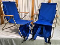 Two blue canvas low folding chairs with metal frames, wooden armrests, and backpack shoulder straps shown fully set up and facing forward.