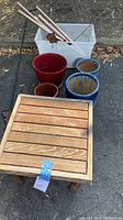 Photo showing the wooden slatted garden table and the collection of four terra-cotta and glazed pots arranged behind it on outdoor pavement.