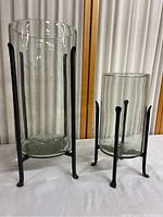 Pair of cylindrical clear glass vases with black metal stands on white surface against curtain background.