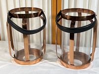 Pair of glass candle holders with copper-toned metal frames and leather handles, placed side by side on a white surface in front of a window with a curtain.
