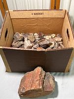 Overall large cardboard box filled with a variety of rock and mineral specimens with one large distinct piece placed in front of the box.