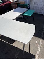 Three folding tables outdoors with two off-white long tables and one smaller green table shown. Tables have folding metal legs and plastic tops.