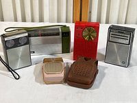 Five portable transistor radios displayed on table: Realistic, Panasonic, red Regency, Westinghouse, pink Global with case