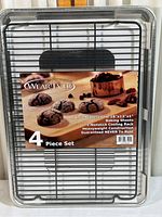 Front view of the set showing three baking sheets stacked together with a nonstick cooling rack on top, all wrapped in original packaging with a label displaying product information and an image of baked goods.