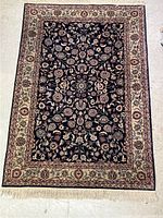 Full view of rectangular wool area rug with floral design on dark field and light border with fringe edges.