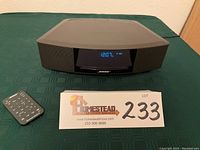 Front view showing Bose unit on green tablecloth, power cord trailing and remote at left with lot ID card.