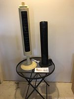 Two tower fans on a black metal mesh foldable patio table. The fans include a taller cream-colored Atlantic Breeze fan and a smaller black Holmes fan.