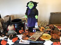 Wide angled shot showing lot of Halloween and autumn decor items on a table including witch figurine, cauldron, pumpkins, baskets, mugs, cookie cutters, flowers, and sign.