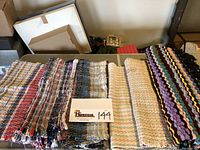 Six assorted handmade rugs or mats stacked and laid out showing variety of colors and patterns.