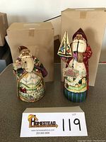 Two Jim Shore Santa figurines displayed with original boxes behind them, one holding a sailing ship and the other with a sheep on a sled. Photo shows full view of both figurines and their distinctive painted details.