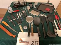 All kitchen utensils, knives, electric sharpener and skillet displayed on green tablecloth.