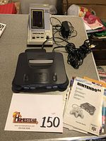 Photo showing Nintendo 64 console, instruction booklets, power cable, and Tandy Space Explorer Zackman device on a table.
