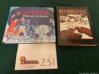Two hardcover coffee table art books side by side on green cloth surface. Left book titled 'Rie Munoz Portrait of Alaska A Thirty-Year Retrospective' with colorful illustrated dust jacket. Right book titled 'The Art of Bev Doolittle' with dust jacket showing snowy winter scene with horses.