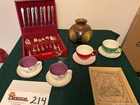 Overall lot on green tablecloth including flatware set in opened red box, four Starbucks cups and saucers, Pottery Craft vase, vintage framed print