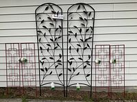 Front view of two tall black metal leaf design trellises and four red geometric zen-style trellises against a white house wall