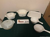Photo showing seven pieces of vintage Corning Ware and Anchor Hocking cookware including casseroles, saucepans, and a tray on a green tablecloth.