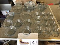 Full lot overview of clear glass bowls, custard cups, baking dishes, refrigerator dish, covered casserole with trivet, mugs, and pie plates set on table.