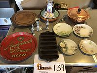 Photo showing full assortment with vintage Muehlebach Pilsner Beer tray, wooden serving plate, pewter salt and pepper shakers with candlestick, covered copper pan, pottery garden girl server, cornbread pan, and four floral plates.