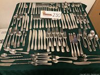Photo shows full assortment of vintage stainless steel flatware laid out on a dark green cloth including knives, forks, spoons, tongs, and serving utensils.